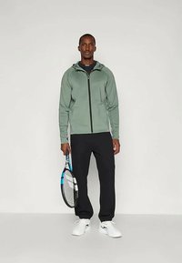 Matšijakk tuhmrohelises värvitoonis, lukuga ees, kombineeritud mustade pükstega. Valged spordijalatsid. Hoides tennise reketit sinise ja musta disainiga.