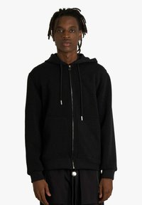 Felpa nera con zip frontale, cappuccio con coulisse e due tasche frontali. La trama del tessuto appare morbida e calda.