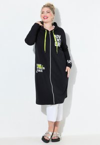 Smiling woman with blonde hair in a black long hoodie dress with neon drawstrings, white cropped pants, and silver sandals against white background.