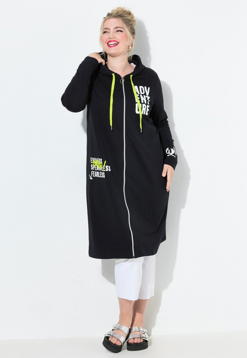 Smiling woman with blonde hair in a black long hoodie dress with neon drawstrings, white cropped pants, and silver sandals against white background.