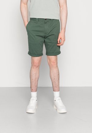Shorts en coton verts avec des ourlets roulés, dotés de poches latérales et d'une fermeture à bouton. Associés à des baskets blanches et des chaussettes blanches à côtes.
