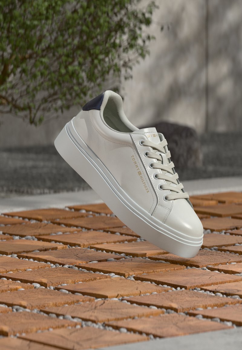 Λευκό sneaker χαμηλού τύπου με κορδόνια και μαύρο ταμπελάκι στη φτέρνα, με το λογότυπο "Tommy Hilfiger", που αιωρείται πάνω από ξύλινη και πλακόστρωτη επιφάνεια σε εξωτερικό χώρο.