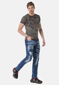 Graues Grafik-T-Shirt mit schwarzem Text auf der Vorderseite, kombiniert mit ausgewaschenen blauen Jeans. Schwarze und rote Sneakers runden das Outfit ab.
