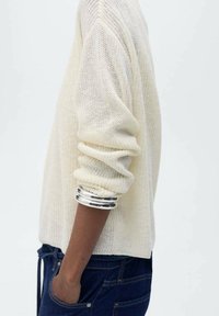 Hellbeige Strickpullover mit gerippten Ärmeln und lockerer Passform. Das Modell trägt silberne Armreifen, kombiniert mit dunklen Jeans.