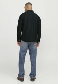 Schwarzes Langarmhemd mit Knopfmanschetten, kombiniert mit lockeren, hellblauen Jeans und braunen Schuhen, von hinten betrachtet.