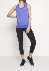 Femme portant un débardeur de sport violet, des leggings noirs coupés et des baskets noires, debout avec une main sur la hanche.