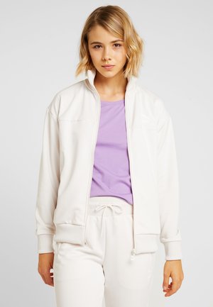 Giacca bianca con zip e polsini a costine, indossata sopra una maglietta viola. Il materiale appare liscio, progettato per l'abbigliamento casual.