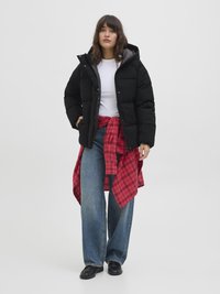 Crna puffer jakna s kapuljačom, velike quilted sekcije, siva podstava, nosi se preko bijele majice, u paru s plavim širokim trapericama i crvenom karirastom košuljom.