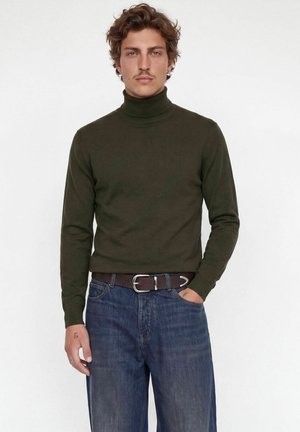 Jeune homme aux cheveux bouclés châtain clair porte un pull à col roulé vert foncé, un jean bleu et une ceinture marron foncé, debout avec une main dans la poche.