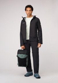 Veste noire résistante à l'eau avec capuche ajustable, associée à un sac à dos noir et vert, un pantalon noir et des chaussures de plein air bleues.