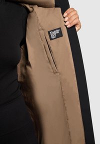 Chaqueta exterior negra con un forro interior marrón claro, que presenta una etiqueta visible y una textura suave. La chaqueta está abierta, mostrando el material interno.