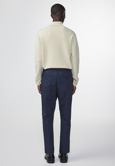 Lichtbeige gebreide trui met hoge kraag, gecombineerd met donkere marineblauwe broek met een elastische tailleband en achterzakken. Zwarte schoenen.