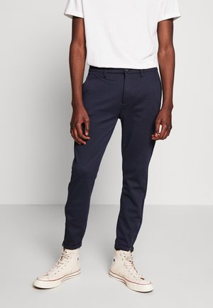 Man in marineblauwe slim-fit broek, witte hoge sneakers en een wit T-shirt met korte mouwen, staand tegen een effen achtergrond.