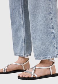 Zilveren metallic sandalen met straps en een vlakke zwarte zool, voorzien van een verstelbare enkelband, gecombineerd met blauwe wijdvallende denimmen.
