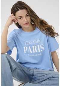 Světle modré bavlněné tričko s bílým potiskem textu "TRÈS CHIC PARIS" a "Toujours à la mode". Volný střih, krátké rukávy, v kombinaci se světle modrými džíny.