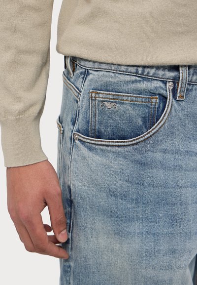 Ανοιχτό μπλε τζιν παντελόνι με φθαρμένη υφή, διαθέτοντας μεταλλικό κουμπί σε ασημί απόχρωση και λεπτομέρειες ραψίματος, ορατό σχέδιο τσέπης και σκούρες ραφές.