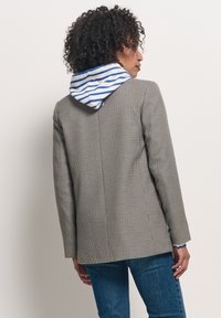 Marynarka w wzór houndstooth w kolorze szarym z kontrastującym niebieskim i białym paskiem na kapturze. Dopasowany krój, gładki materiał, pojedyncze rozcięcie z tyłu.