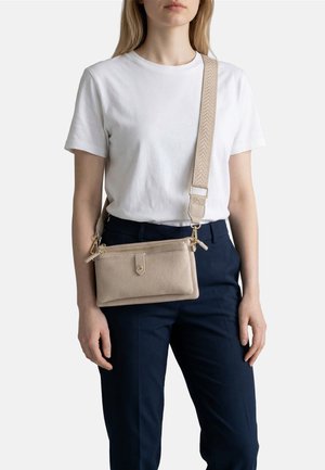 Donna con maglietta bianca e pantaloni blu navy, con una borsa a tracolla beige caratterizzata da una tracolla larga con motivo e scomparti con zip.