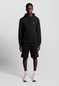 Felpa nera con logo giallo, abbinata a pantaloni corti neri. Il tessuto appare liscio e il design presenta una tasca a marsupio e cordini.