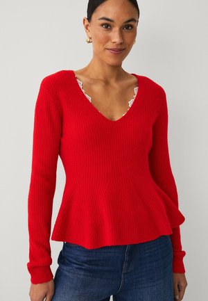 Woman wearing a red ribbed sweater with peplum hem and lace-trimmed white top underneath, paired with dark blue jeans.