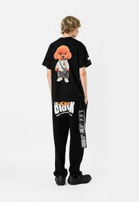 Camiseta de algodón negra con un gráfico de un poodle de dibujos animados en la parte trasera, combinada con pantalones de chándal negros con texto y patrones en blanco. Calzado negro.