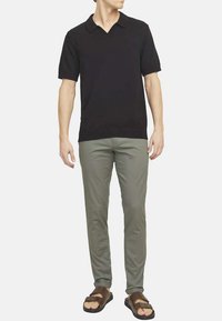 Polo shirt noir à manches courtes, en tissu tricoté, associé à un pantalon slim vert clair et des sandales marron, se tenant sur un fond neutre.