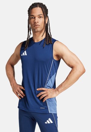Homme avec de longues dreadlocks portant un débardeur de sport bleu et un short assorti, debout les mains sur les hanches devant un fond uni.
