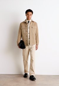 Veste en daim tan avec deux poches avant sur une chemise crème, pantalon beige et chaussures noires. Tenant une pochette noire, debout contre un mur blanc.