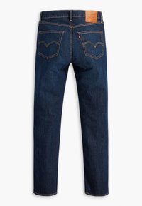 Jean en denim bleu foncé vu de dos avec deux poches arrière cousues, un patch de marque en cuir marron et une étiquette rouge sur la poche droite.