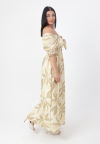 Robe maxi en tissu crème léger avec de grands motifs floraux verts et beiges. Présente des manches dénudées et un design smocké à la taille.
