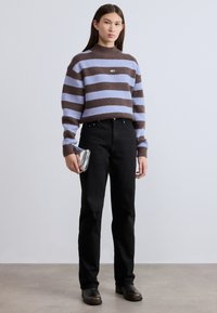 Πλεκτό πουλόβερ με ρίγες σε λιλά και καφέ, με ψηλό λαιμό, συνδυασμένο με μαύρο τζιν σε ίσια γραμμή και μαύρα μποτάκια μέχρι τον αστράγαλο, κρατώντας μία ασημί τσάντα φάκελο.