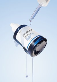 Szklana butelka z zakraplaczem o niebieskim gradientowym odcieniu, białą nakrętką i etykietą. Przezroczyste serum kapie z zakraplacza i butelki, na tle jasnoniebieskiego tła.