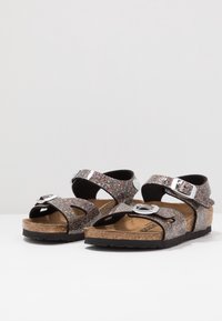 Błyszczące czarne sandały z korkową wkładką i regulowanymi srebrnymi klamrami, z wieloma wycięciami na paskach oraz fakturowaną czarną podeszwą.