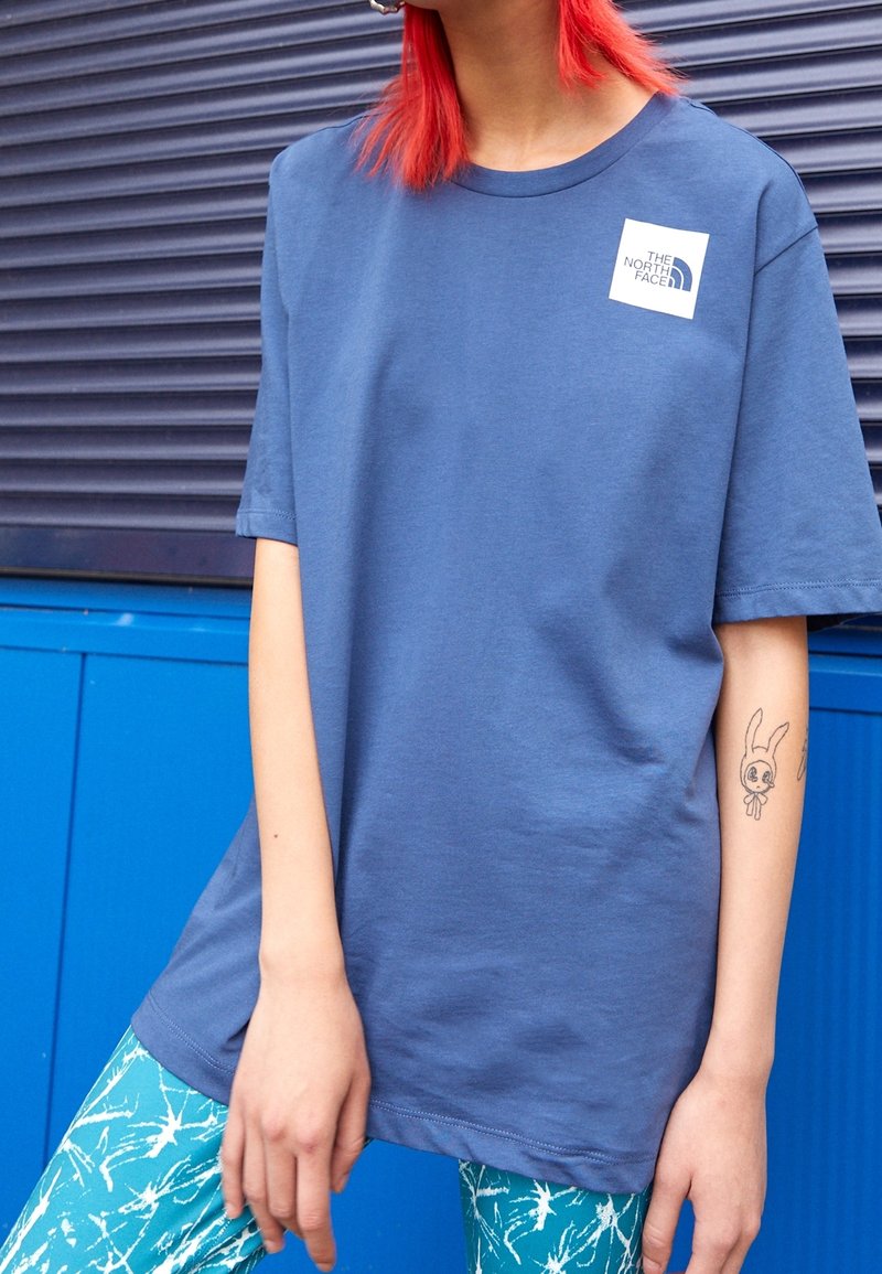 T-shirt en coton bleu à manches courtes avec un patch logo sur la poitrine. La personne porte des leggings verts à motifs et un tatouage visible sur le bras.