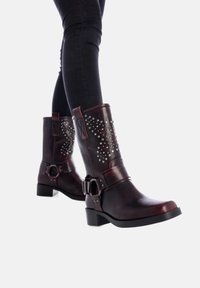 Botas de cuero marrón con tachuelas decorativas, hebillas laterales, un tacón grueso y un acabado suave, sobre jeans skinny negros.