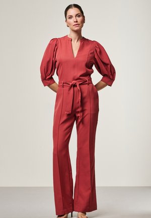 Frau steht mit den Händen hinter dem Rücken, trägt einen rostrot-gürtelten Jumpsuit mit Puffärmeln und High Heels, vor einem einfarbigen Hintergrund.