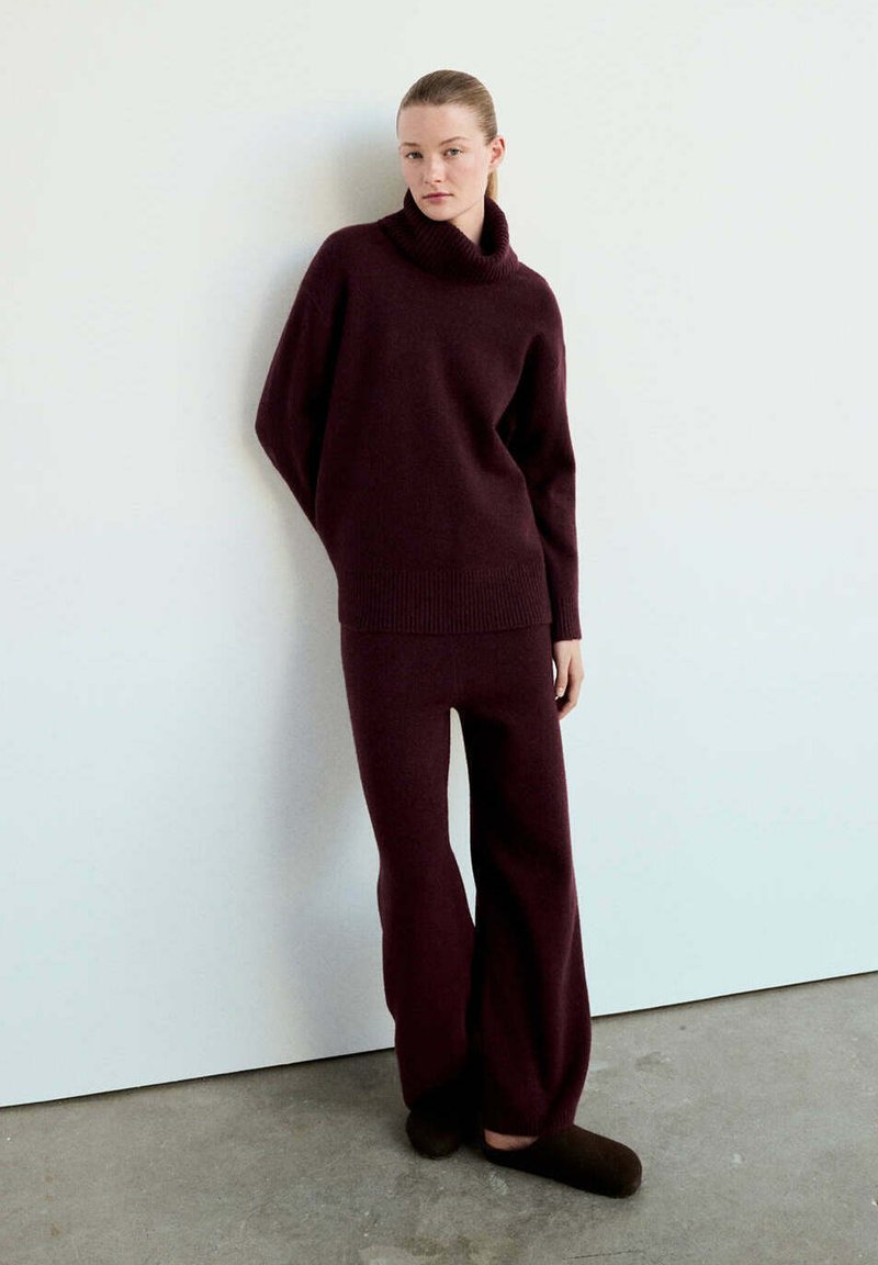 Femme debout contre un mur blanc, portant un pull à col roulé violet foncé, un pantalon large assorti et des chaussons foncés sur un sol en béton.