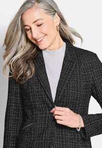 Blazer texturé en noir et blanc avec une coupe structurée, présentant un col à revers et des poches à zip, superposé sur un haut gris clair.