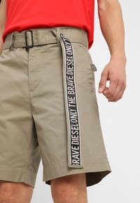 Pantaloni corti khaki con una cintura laterale in tessuto a rete che presenta un logo ripetuto in nero, bianco e rosso. Il tessuto è liscio e con un design strutturato.