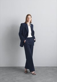 Námořnický modrý oblekový sako a kalhoty, spárované s bílou košilí. Slim fit, hladká textura a černé sandály s otevřenou špičkou.