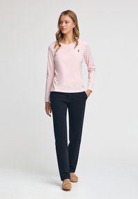 Ljusrosa långärmad tröja med rund halsringning och en liten mörk logotyp. Matchas med mörka byxor och beigea slip-on-skor.