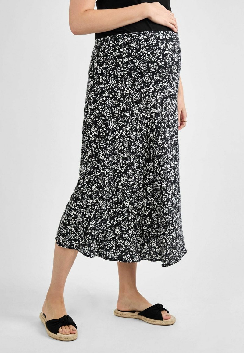 Falda de maternidad negra con estampado floral en blanco, diseño fluido, longitud hasta la mitad de la pantorrilla, textura suave, combinada con sandalias de tiras negras con nudo.