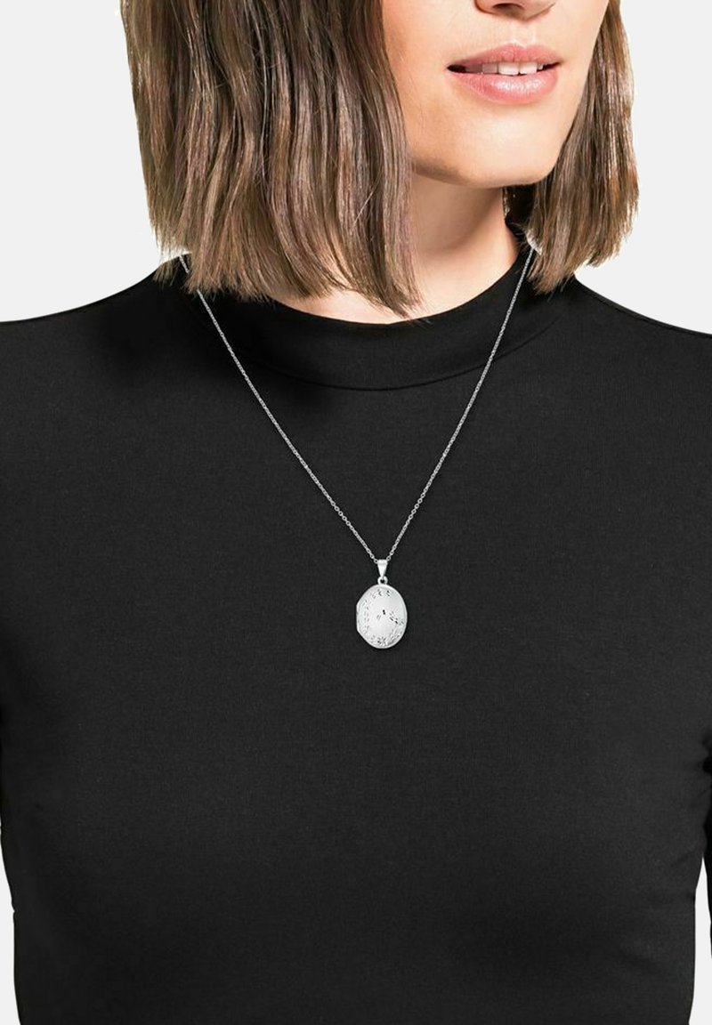 Silverhalsband med en oval form och intrikata blommönster, hängande på ett delikat kedja, bärs över en svart topp.