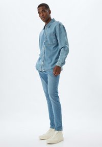 Giacca di jeans azzurro chiaro con una singola tasca sul petto, abbinata a jeans azzurro chiaro a taglio slim. Modello che indossa sneakers bianche alte.