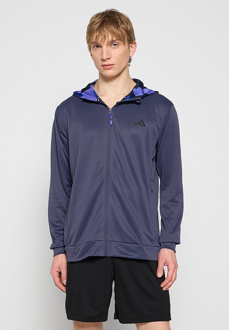 Veste de sport navy avec zip sur le devant, doublure de capuche violette contrastante, deux poches latérales et le logo Adidas sur la poitrine. Tissu léger.