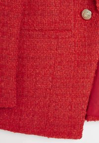 Tissu rouge texturé avec le contour d'une poche et un seul bouton doré orné sur le côté supérieur droit.