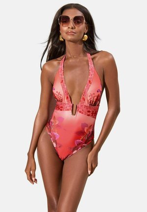 Kobieta w koralowym kwiatowym jednoczęściowym kostiumie kąpielowym z głębokim dekoltem w kształcie litery V, złotymi kolczykami i dużymi okularami przeciwsłonecznymi, stojąca na jednolitym tle.