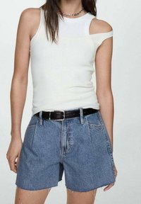 Top de tirantes blanco con un hombro descubierto, combinado con pantalones cortos de denim azul, con un cinturón negro y dobladillos deshilachados.