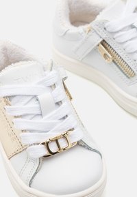 Sneakers in ecopelle bianca con dettagli in cerniera dorata e fibbia, dotati di fodera interna texturizzata e lacci piatti bianchi.