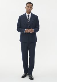 Man står klädd i en marinblå randig dubbelknäppt kostym, vit skjorta, randig slips och svarta finskor på en vit bakgrund.